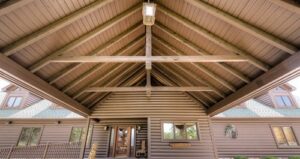 View looking up at the pitched wooden roof of a log cabin entrance, showing exposed beams and a hanging lantern, with windows and doors visible below.