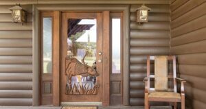 Wooden front door with glass panels and a carved jackrabbit design. Two lantern-style lights are mounted on the log cabin wall, and a wooden chair with a cushioned seat sits to the right of the door.