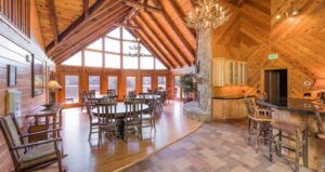 Spacious lodge interior with wood-paneled walls, large windows, round dining tables, rustic chairs, stone fireplace, and antler chandelier under a vaulted ceiling.