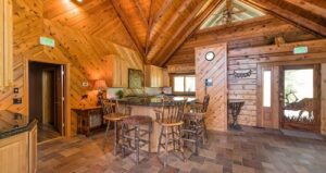 A cozy wooden cabin kitchen with a vaulted ceiling, wooden cabinets, granite countertops, a central island with four stools, and rustic decor. Natural light enters through windows and a glass front door.