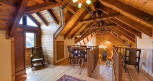 Loft with wood paneling, exposed beams, a table with chairs, rocking chairs, and a view overlooking a lower level with large windows letting in natural light.