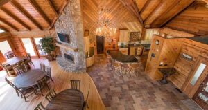 Spacious, rustic cabin interior with wooden walls and ceiling, stone fireplace, chandelier, kitchen with bar stools, round dining tables, and large windows letting in natural light.