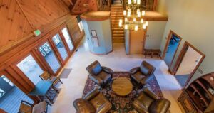 Cozy lodge lobby with four brown leather chairs around a round table on a rug, wooden walls, a chandelier, doors to the outside, staircase, and open doorways leading to other rooms.