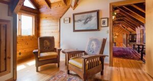 Two wooden armchairs with patterned cushions sit in a cozy, wood-paneled loft. Sunlight streams through a window. Art hangs on the wall, and a hallway with a rug leads to another wooden room.