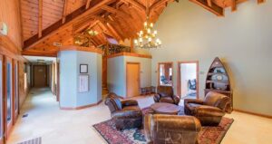 Spacious lodge-style room with wood ceilings, a chandelier, four leather chairs around a round table, area rug, pale walls, and a boat-shaped shelf with decor. Multiple doorways lead to other rooms.