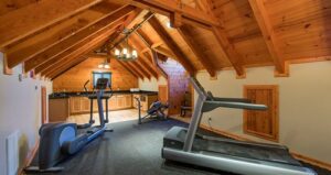 A home gym in a wood-paneled attic features a treadmill, exercise bike, and rowing machine on black carpet, with a kitchenette and chandelier in the background.