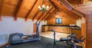A home gym with an elliptical machine and weight bench is set under a vaulted wooden ceiling. A kitchenette with cabinets and a sink is in the background, with wooden floors and a small window above the counter.