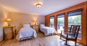 A cozy bedroom with two single beds, wooden furniture, a rocking chair, large windows, and a view of trees and a lake outside. Warm lighting creates a welcoming atmosphere.