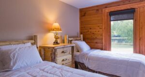 Cozy bedroom with two twin beds, white bedding, a rustic wooden nightstand with a lamp, and a large window overlooking greenery outside.
