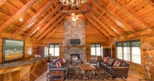 Spacious log cabin living room with vaulted wood ceiling, stone fireplace, leather sofas, a wooden coffee table, and large windows letting in natural light.