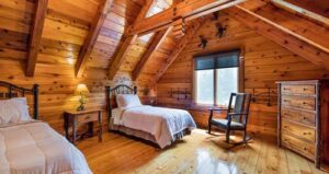 Cozy log cabin bedroom with wood walls and ceiling, two single beds, a rocking chair, dresser, and two lamps. Sunlight streams in through a window and ceiling skylight.