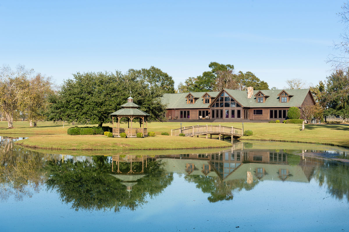 A large wooden house with dormer windows sits behind a pond, with a small bridge and a gazebo in the foreground. Trees and blue sky are reflected in the calm water.