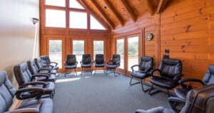 A meeting room with a wood-paneled interior, large windows, vaulted ceiling, and black office chairs arranged in a circle on a carpeted floor. Sunlight streams in through the windows.
