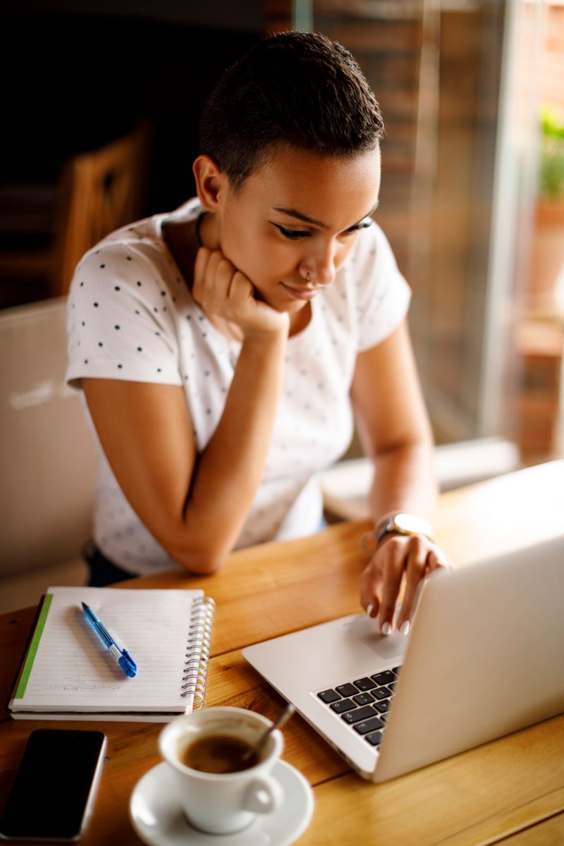 A person sits at a wooden table, using a laptop. There is a notebook with a pen, a smartphone, and a cup of coffee on the table. The person appears focused and is wearing a white polka-dotted shirt.
