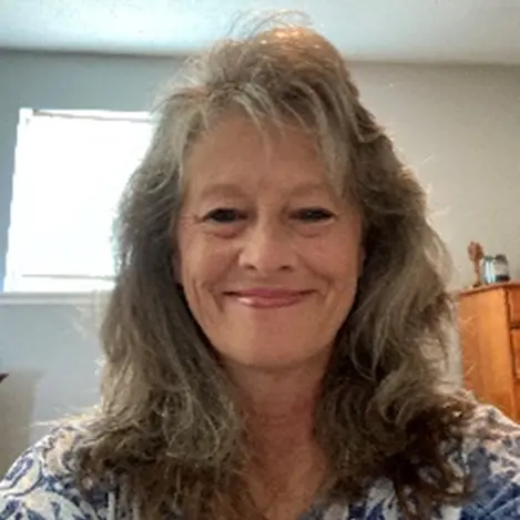 A woman with long, wavy gray hair smiles at the camera in a well-lit room with a light-colored wall and window in the background.