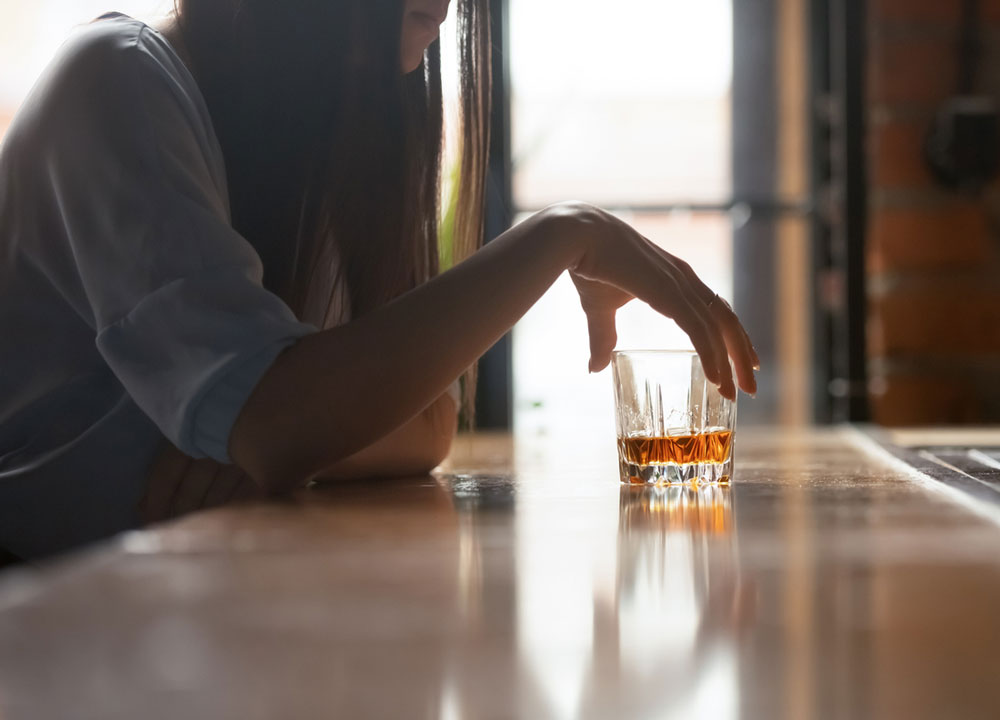A person with long hair sits at a wooden bar, resting their arm on the counter and holding a glass of whiskey. The scene is softly lit, creating a contemplative mood.