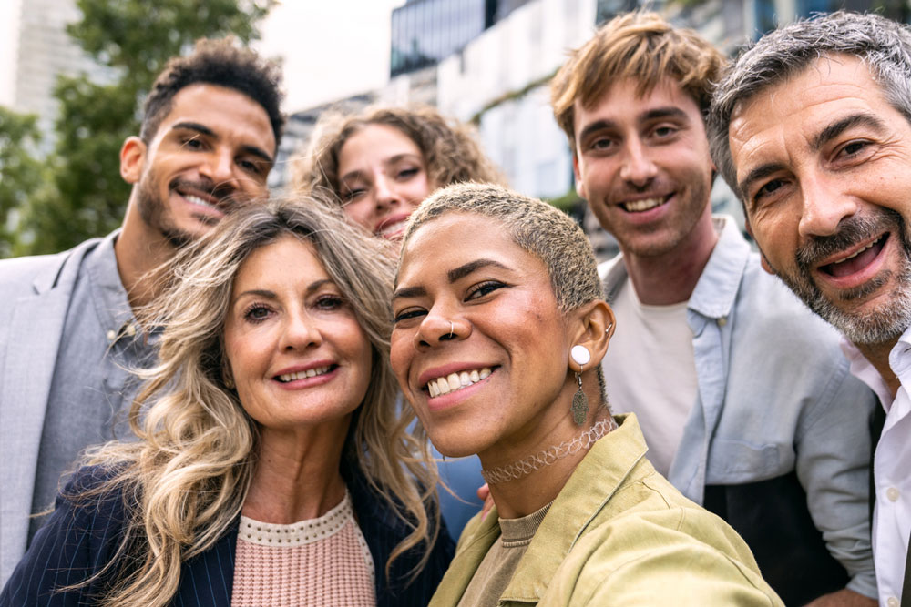 Six adults of diverse backgrounds stand close together outdoors, smiling and posing for a group selfie on a sunny day, with buildings and trees in the blurred background.