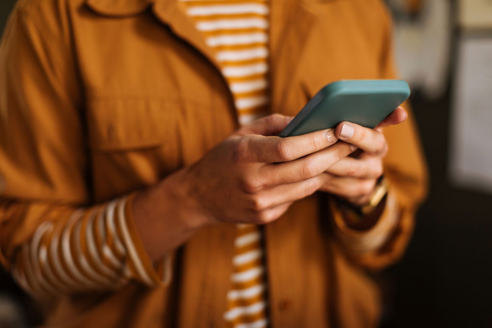 A person wearing a brown shirt and striped top holds a smartphone, using both hands. The image is focused on the hands and phone, with the face out of frame.
