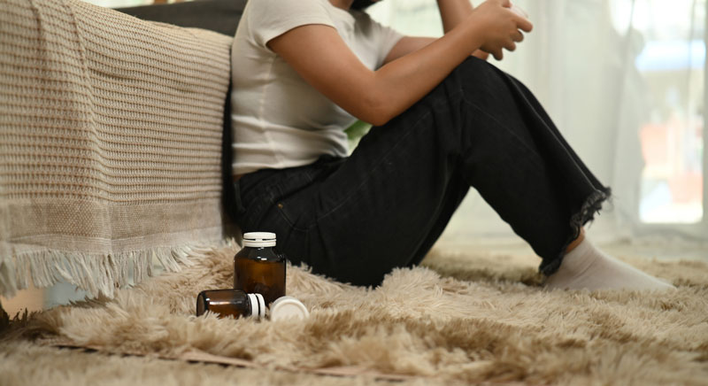 A person sits on a shaggy rug next to a couch, with two pill bottles—one open—lying on the floor nearby. The persons face is not visible.