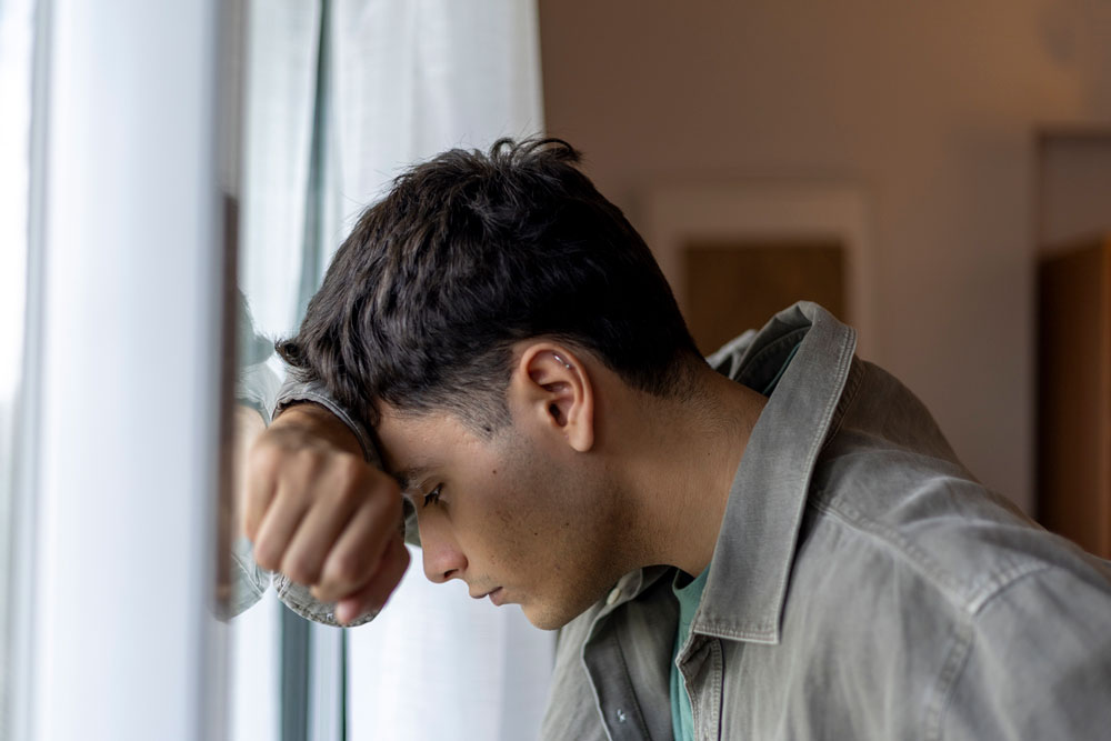 Young man with short dark hair leans his forehead and arm against a window, looking down with a serious expression, wearing a gray jacket indoors.
