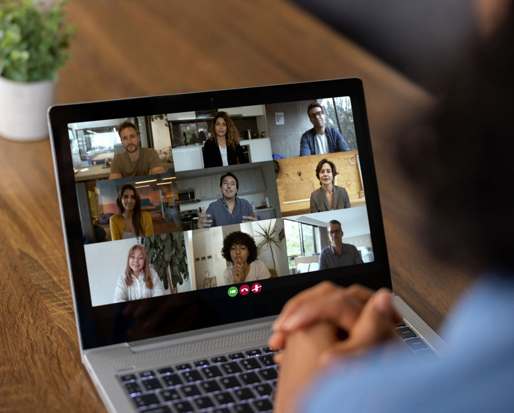 A person participates in a video call on a laptop with eight people visible in small windows, each in different indoor settings, suggesting a virtual meeting or group discussion.