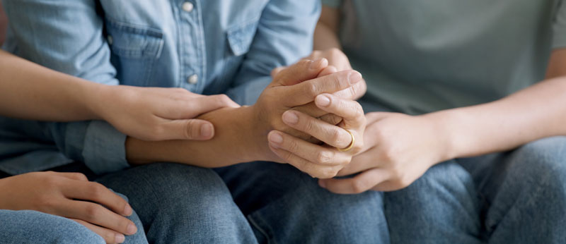 Three people sit closely together, holding and comforting each others hands. They wear casual clothes, and the scene suggests support, care, and empathy in a difficult moment.