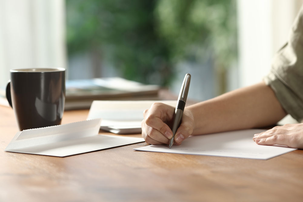 A person writes a letter on a wooden table beside a black mug and an open envelope, with blurred greenery visible in the background.