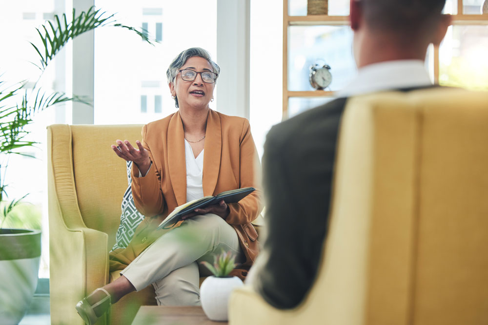 A professional woman with short gray hair and glasses speaks to a man in an office, holding a notebook and gesturing with her hand while seated in a yellow chair.