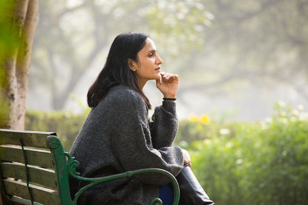A woman sits on a green park bench, wearing a gray sweater, looking thoughtful. The background is blurred greenery and trees.