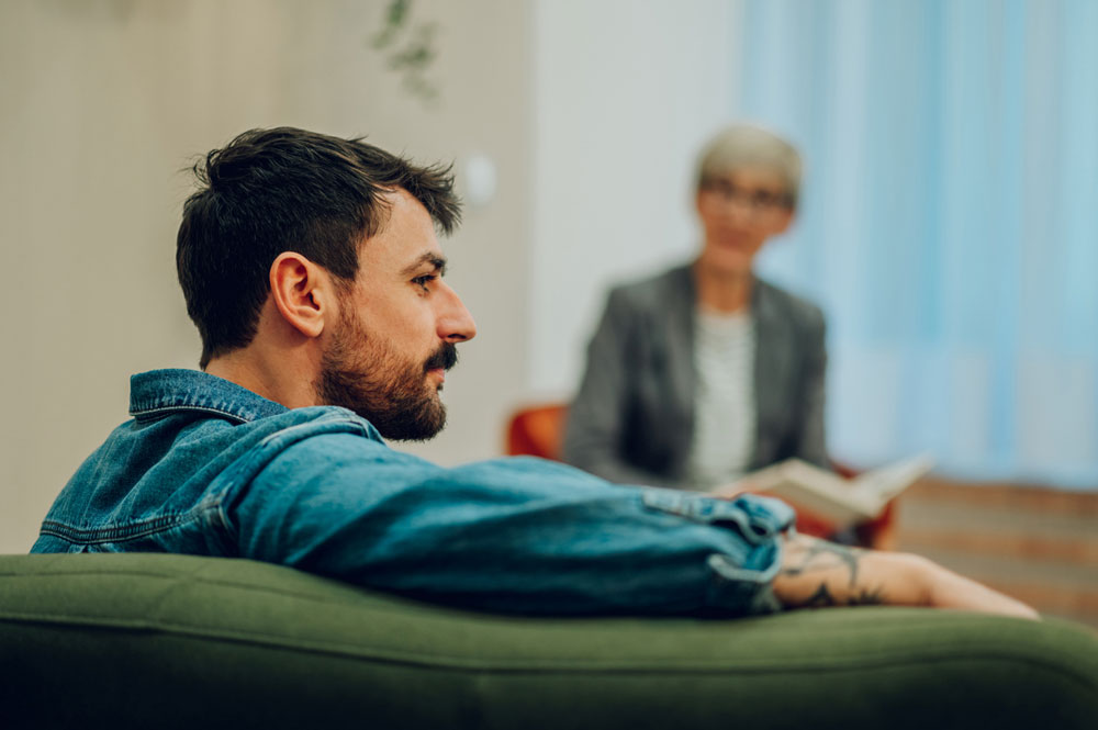 A man with a beard sits on a green couch, looking thoughtful. In the background, an older person with glasses and a notebook sits, slightly out of focus, suggesting a counseling or therapy session.