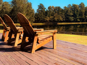 Two wooden Adirondack chairs on a deck overlook a calm lake, surrounded by green grass and trees under a clear blue sky.