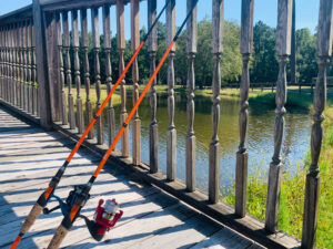 Two fishing rods rest against a wooden bridge railing, with a pond and green trees visible in the background on a sunny day.
