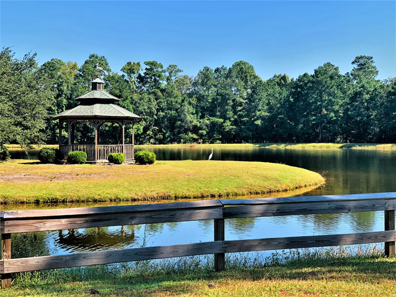 A wooden gazebo sits by a pond surrounded by grass and trees, with a wooden fence in the foreground and a white bird standing by the water on a sunny day.