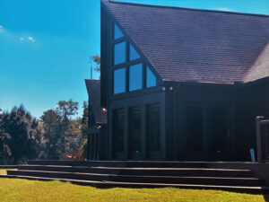 Modern cabin-style house with large triangular windows and dark exterior, surrounded by grass and trees, under a bright blue sky.