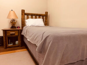A neatly made twin bed with a wooden headboard, gray blanket, and white pillow is next to a wooden nightstand holding a lamp, clock, and books in a simple, beige-walled room.