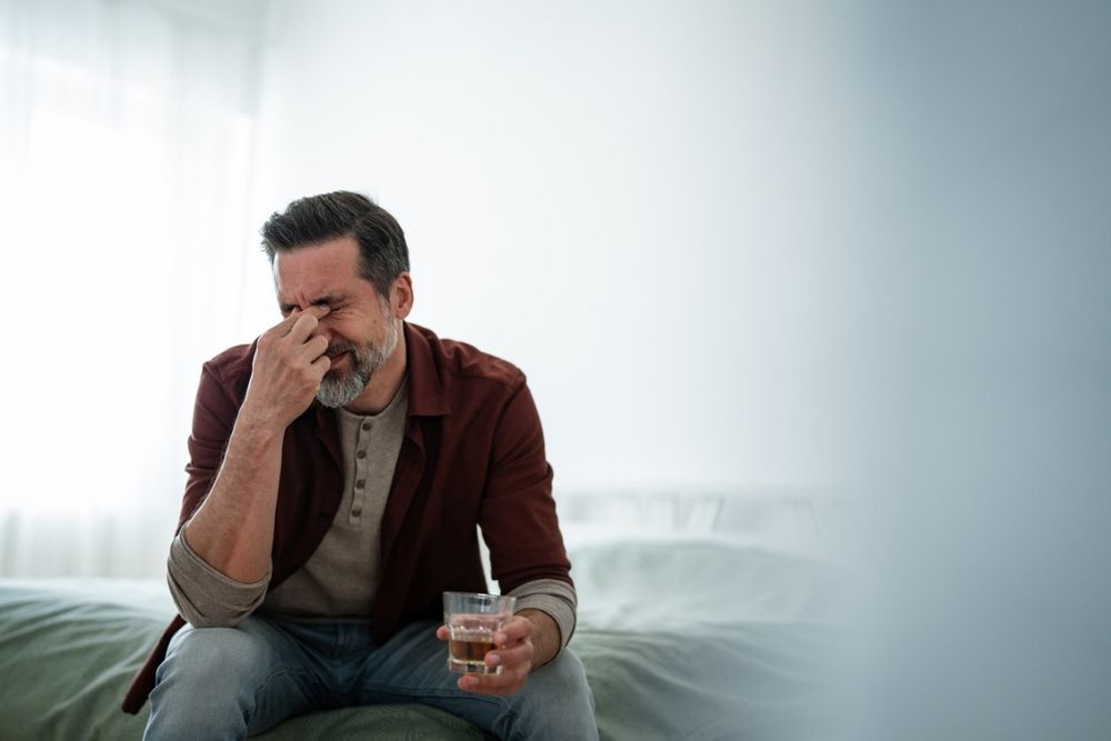 A man sits on a bed, pinching the bridge of his nose with his eyes closed, looking stressed. He holds a glass of water in his other hand. The background is softly lit and out of focus.