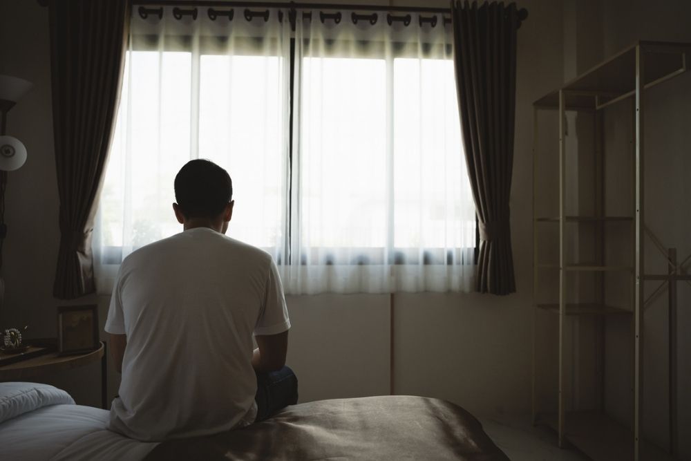 A person sits on the edge of a bed facing a window with sheer curtains, in a dimly lit bedroom. The person is silhouetted and appears to be deep in thought.