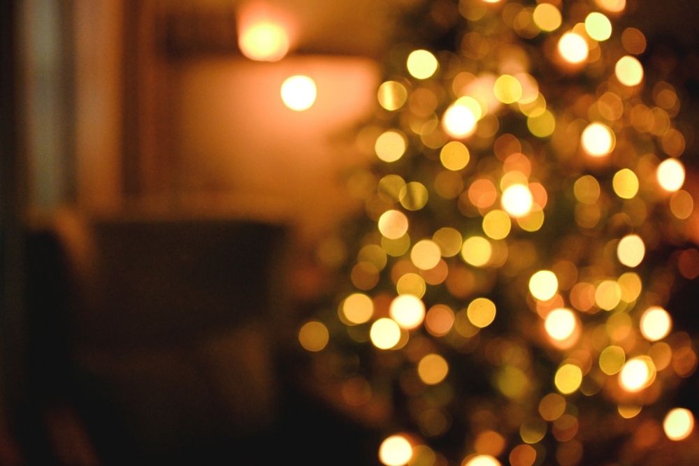 Out-of-focus image of warm yellow and orange lights, likely holiday string lights on a tree, creating a soft, festive bokeh effect. The background is dark, with indistinct furniture shapes visible.