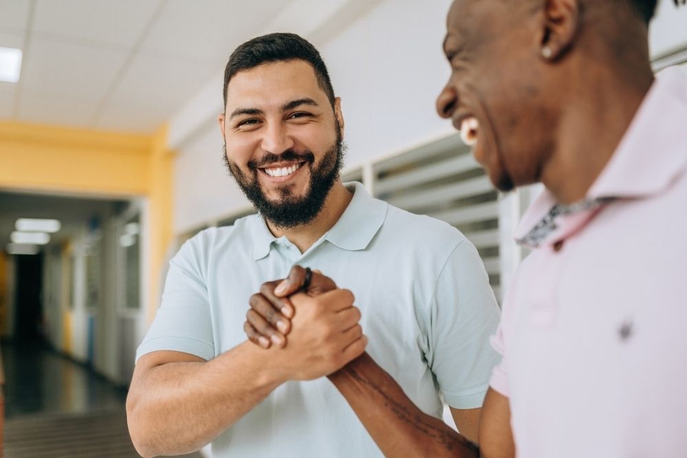 Two men smiling and clasping hands in a friendly greeting inside a bright hallway. Both appear happy and relaxed, with one man facing the camera and the other in profile.