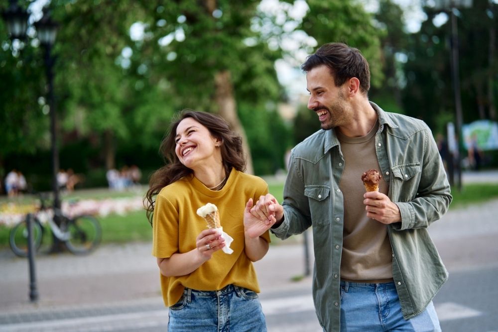 A smiling couple walks outside holding hands and eating ice cream cones. They look happy, dressed casually in jeans and shirts, with greenery and a bike in the background.