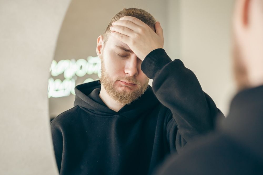 A person wearing a black hoodie stands in front of a mirror with their hand on their forehead, eyes closed, appearing stressed or tired.