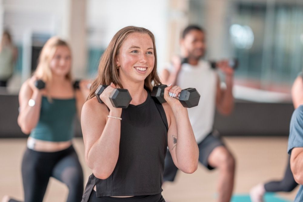 A group of people exercise indoors, holding dumbbells and performing lunges. They appear focused and are wearing athletic clothing. The setting is bright and modern.
