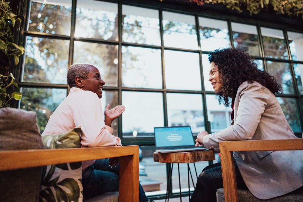 Two people sit across from each other at a small table in a cozy, plant-filled room, talking and smiling. A laptop is open between them, and large windows in the background let in natural light.