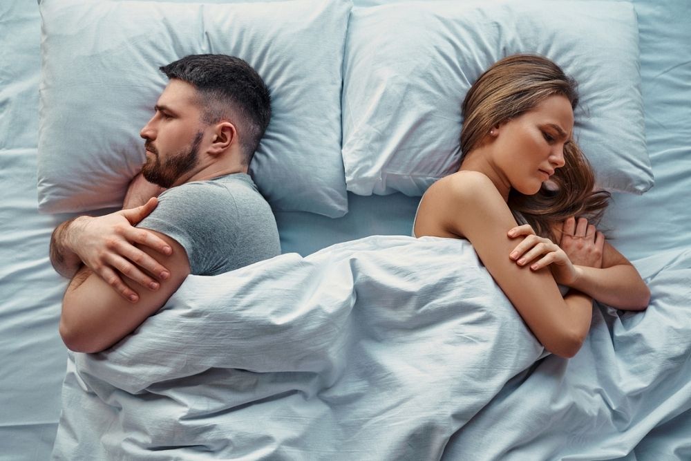 A man and woman lie in bed, turned away from each other with arms crossed, both appearing upset or distant.