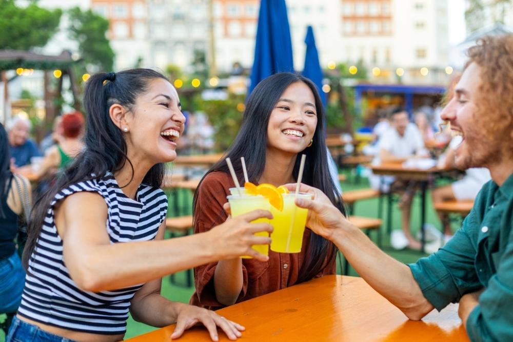 Three friends sit at an outdoor table, smiling and clinking glasses filled with yellow drinks. Other people and string lights are visible in the background, creating a lively, festive atmosphere.