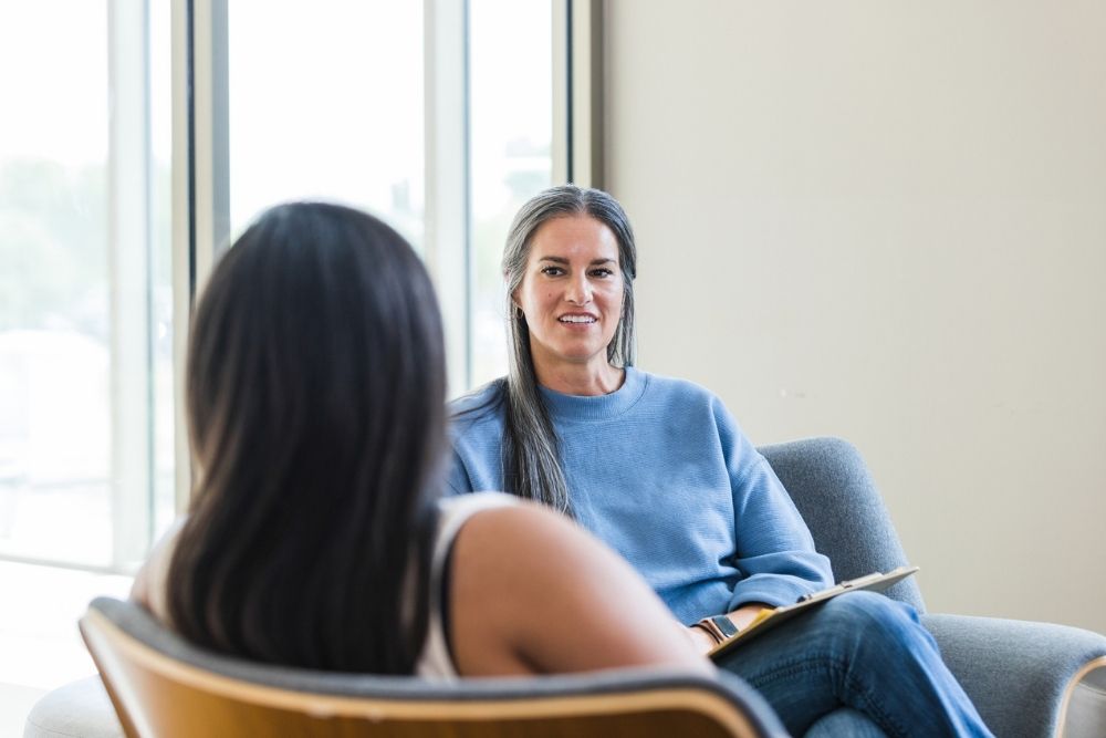 Two women sit facing each other indoors. One woman with long gray hair and a blue sweater is smiling, holding a notebook, while the other woman has her back to the camera.