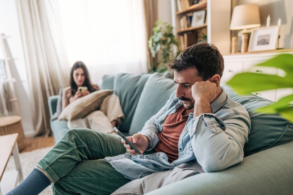 A man sits on a couch looking at his phone, appearing thoughtful or concerned. In the background, a woman sits on another couch, also focused on her phone. The room looks cozy and well-lit.