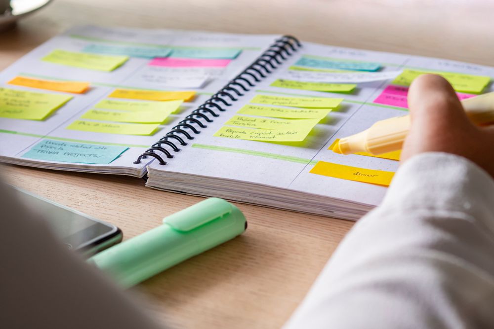 A person highlights notes in a spiral notebook filled with colorful sticky notes and organized sections. A green highlighter and a smartphone lie nearby on the desk.