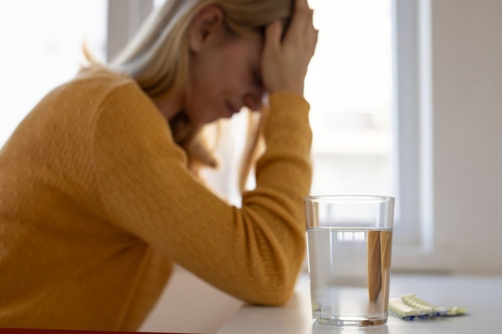 A woman in a yellow sweater sits with her head in her hand, looking stressed. A glass of water and some pills are on the table in front of her, with a window in the background.