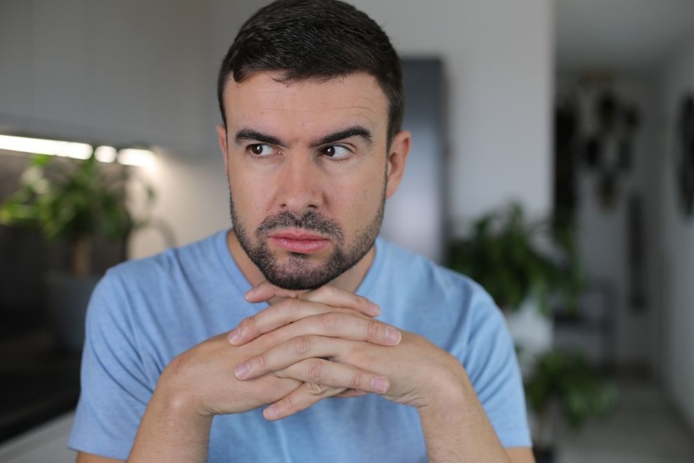 A man with short dark hair and a beard sits indoors, resting his chin on his hands and looking to the side with a thoughtful, slightly concerned expression. There are green plants in the blurred background.